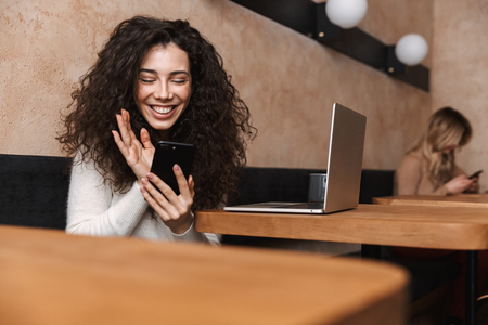 Image of pretty happy girl sitting in cafe using laptop computer and mobile phone waving.の写真素材