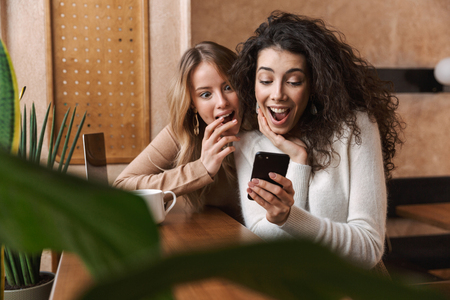 Two cheerful young girlfriends sitting at the cafe indoors, having cup of coffee, using mobile phone, chattingの写真素材