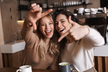 Two cheerful young girls friends sitting at the cafe table, having fun together, drinking coffeeの写真素材