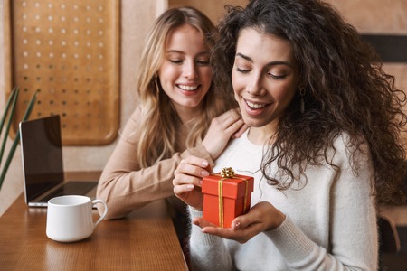 Two cheerful young girlfriends sitting at the cafe indoors, having cup of coffee, holding present...