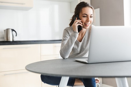Attractive smiling young woman working on laptop computer while sitting on a kitchen, using mobile phoneの写真素材