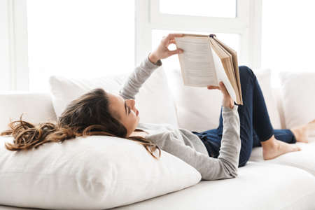 Smiling beautiful girl relaxing on a couch at home, reading a book