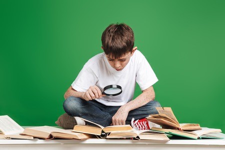 Cute little boy with freckles studying, sitting with stack of books over green