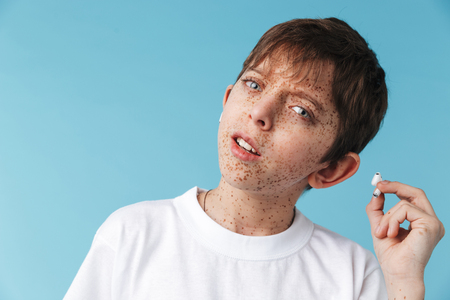 Image of happy caucasian boy 10-12y with freckles wearing white casual t-shirt and earpods looking at camera isolated over blueの写真素材