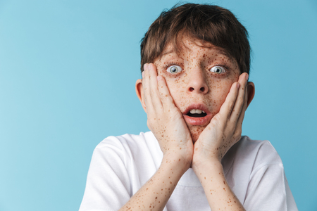 Image closeup of excited caucasian boy 10-12y with freckles wearing white casual t-shirt smiling at camera isolated over blueの写真素材