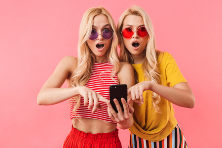 Shocked blonde twins in sunglasses using smartphone and looking at the camera over pink backgroundの写真素材