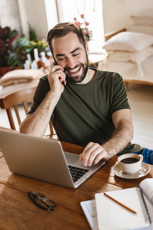 Photo of unshaved mature man 30s in casual clothing using laptop and smartphone while working in flatの写真素材