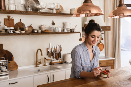 Photo of gorgeous brunette woman 20s eating panna cotta dessert with teaspoon while having breakfast in stylish kitchen at homeの写真素材