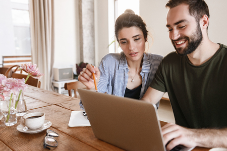 Image of beautiful brunette couple man and woman 20s drinking coffee and working on laptop together while sitting at table at homeの写真素材