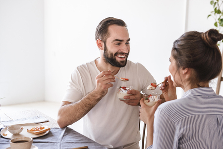 Image of joyous brunette couple man and woman 20s eating panna cotta dessert together while sitting at table at homeの写真素材