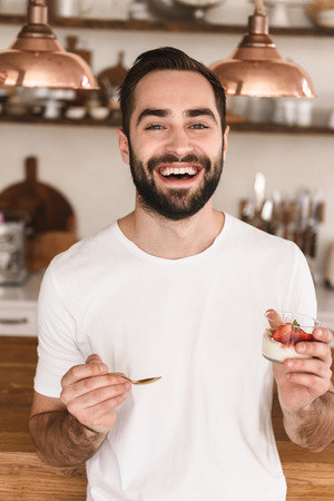 Portrait of joyful brunette man 20s eating panna cotta dessert with teaspoon while having breakfast in stylish kitchen at homeの写真素材