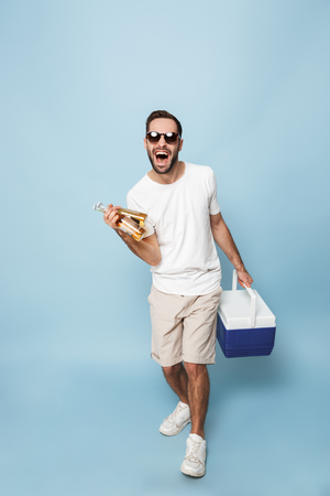 Full length of a cheerful excited man wearing blank t-shirt standing isolated over blue background, carrying cooler with cold beerの写真素材