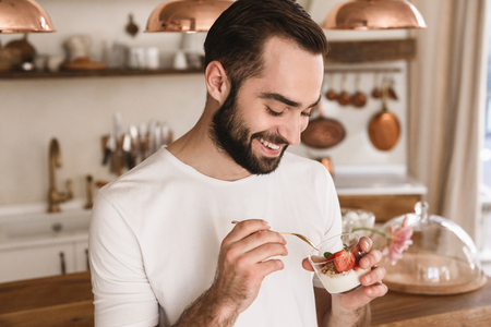 Portrait of positive brunette man 20s eating panna cotta dessert with teaspoon while having breakfast in stylish kitchen at homeの写真素材
