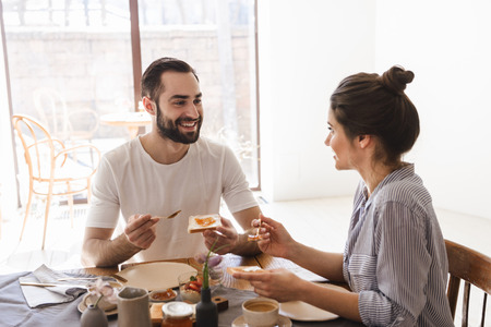 Image of satisfied brunette couple man and woman 20s eating breakfast together while sitting at table in apartmentの写真素材