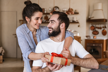 Photo of happy brunette couple man and woman 20s having breakfast in apartment while sitting at table with present boxの写真素材