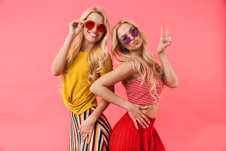 Pleased blonde twins in sunglasses posing together and looking at the camera over pink backgroundの写真素材