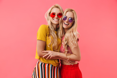 Happy blonde twins in sunglasses hugging and looking at the camera over pink backgroundの写真素材