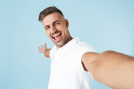 Image of a handsome excited emotional adult man posing isolated over blue wall background take a selfie by camera.の写真素材