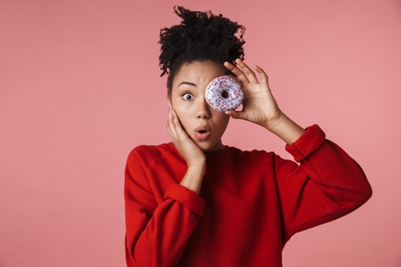 Image of a beautiful amazing happy excited young african woman posing isolated over pink wall background holding sweeties donut.の写真素材