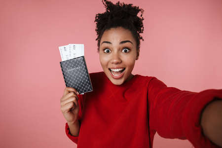 Image of a beautiful amazing happy excited young african woman posing isolated over pink wall background take selfie by camera with passport and tickets.の写真素材
