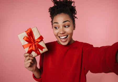 Image of a beautiful amazing happy excited young african woman posing isolated over pink wall background take selfie by camera with present gift box.の写真素材