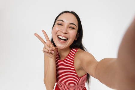 Photo of a beautiful young pretty asian excited woman posing isolated over white wall background take selfie by camera.の写真素材