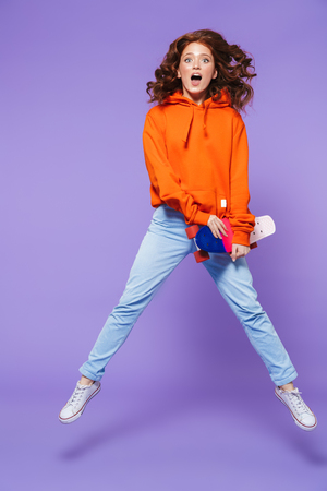 Full length portrait of a pretty young redheaded woman jumping isolated over violet background, holding skateboardの写真素材