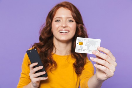 Portrait of a pretty cheerful young redheaded woman standing isolated over violet background, using mobile phone, showing credit cardの写真素材