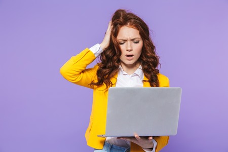 Portrait of a pretty shocked young redheaded woman standing isolated over violet background, using laptopの写真素材