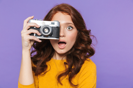 Photo of positive redhead woman wearing yellow clothes holding retro vintage camera and taking picture isolated over purple backgroundの写真素材