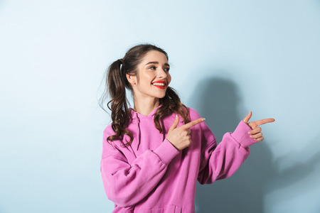 Portrait of stylish young woman with two ponytails smiling and pointing fingers at copyspace isolated over blue background in studioの写真素材