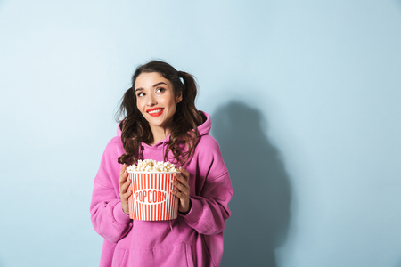 Portrait of pretty young woman with two ponytails smiling while holding popcorn bucket isolated over blue background in studioの写真素材