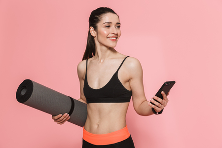 Image of amazing beautiful young pretty fitness woman using mobile phone holding rug isolated over pink wall background.の写真素材