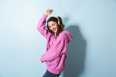 Portrait of positive young woman wearing headphones listening to music and dancing isolated over blue background in studioの写真素材