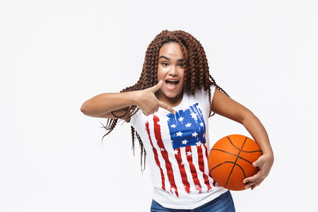Portrait of attractive african american woman holding basketball during game while standing isolated against white wallの写真素材