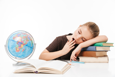 Portrait of sleepy school girl reading studying books and doing homework while sitting at desk in class isolated over white backgroundの写真素材