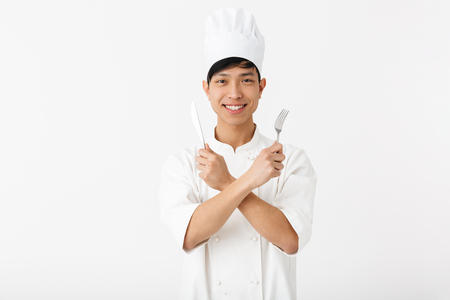 Image of asian positive chief man in white cook uniform smiling at camera while holding cutlery isolated over white backgroundの写真素材