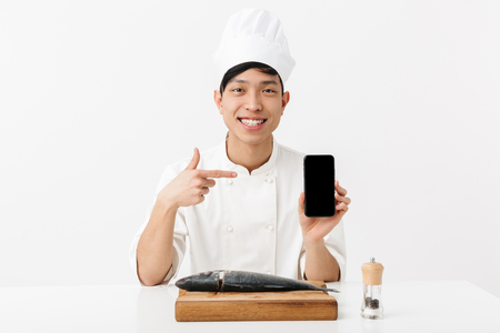 Image of joyous japanese chief man in white cook uniform holding smartphone while fillet raw fresh fish isolated over white backgroundの写真素材