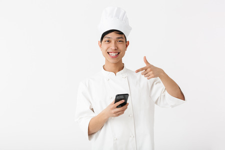 Image of positive chinese chief man in white cook uniform and chef's hat holding mobile phone isolated over white backgroundの写真素材
