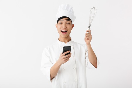 Excited asian chef wearing uniform standing isolated over white background, using mobile phone, showing whiskの写真素材