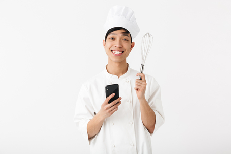 Excited asian chef wearing uniform standing isolated over white background, using mobile phone, showing whiskの写真素材