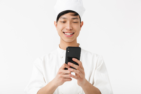 Image of cheerful chinese chief man in white cook uniform and chef's hat holding mobile phone isolated over white backgroundの写真素材