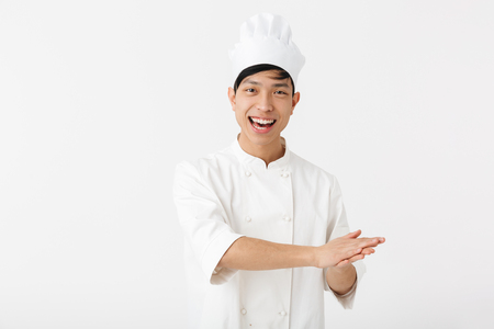 Portrait of happy chinese man in white cook uniform and chef's hat smiling at camera while standing isolated over white backgroundの写真素材