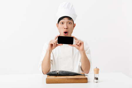 Image of surprised japanese chief man in white cook uniform holding smartphone while fillet raw fresh fish isolated over white backgroundの写真素材