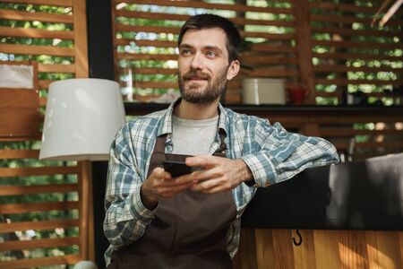 Image of european waiter boy wearing apron sitting at the bar and using cellphone while working in...