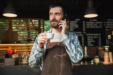 Portrait of handsome barista guy wearing apron drinking coffee and talking on cellphone in street...