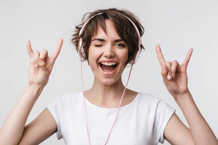 Image of attractive woman in basic t-shirt showing rock sign while listening to music with headphones isolated over white backgroundの写真素材