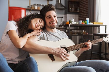 Image of happy young couple man and woman 30s sitting on sofa at home and playing music on acoustic...