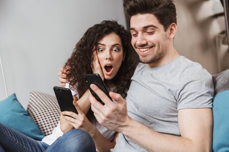Picture of caucasian young couple man and woman 30s sitting on sofa at home and using smartphones togetherの写真素材