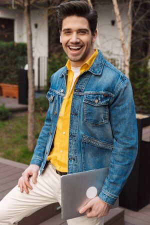 Image of a handsome happy cheerful young business man near cafe outdoors holding laptop computer.の写真素材